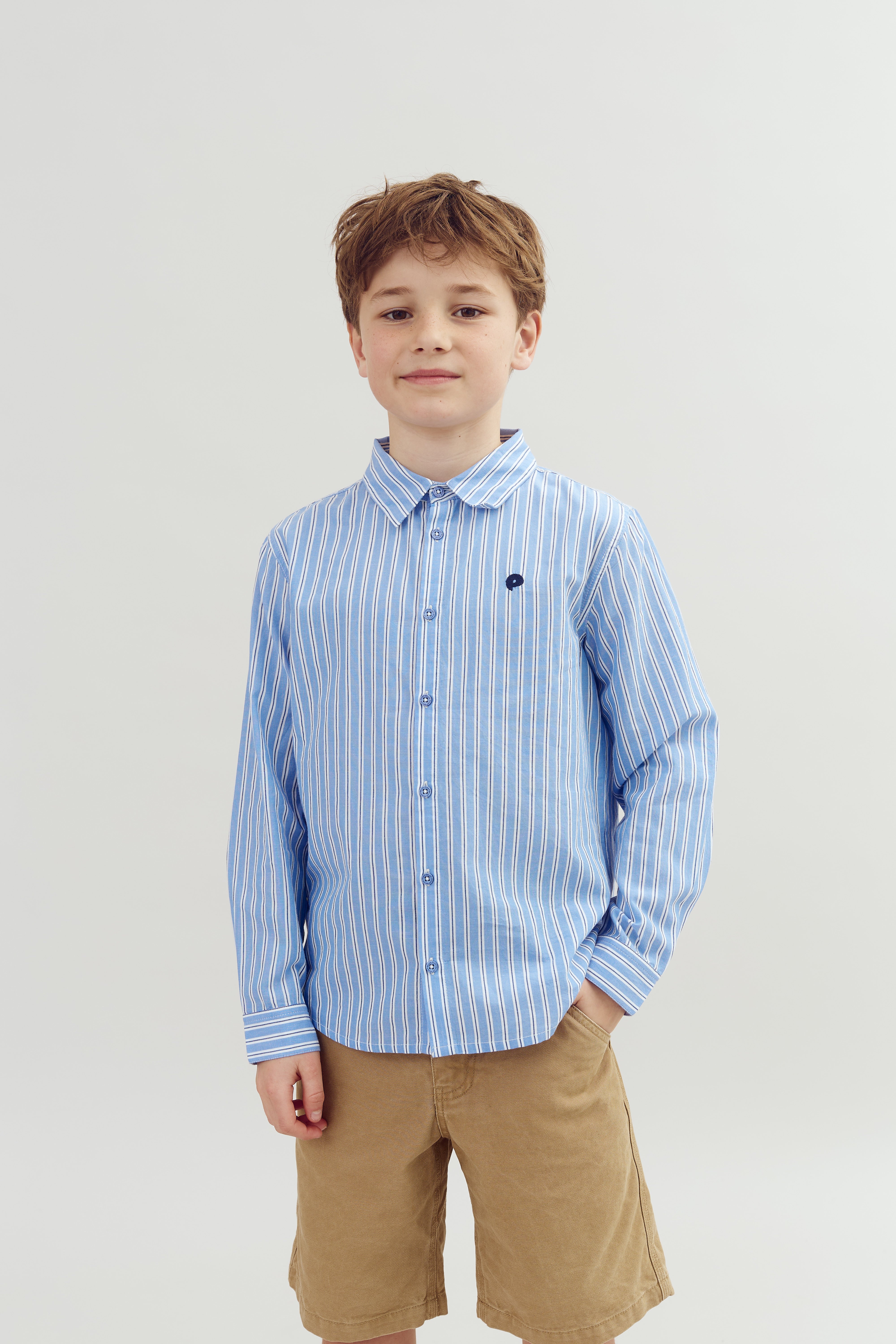 A boy stands, wearing a blue and white striped shirt and tan shorts, in front of a plain light gray background. His left hand rests in his shorts pocket.