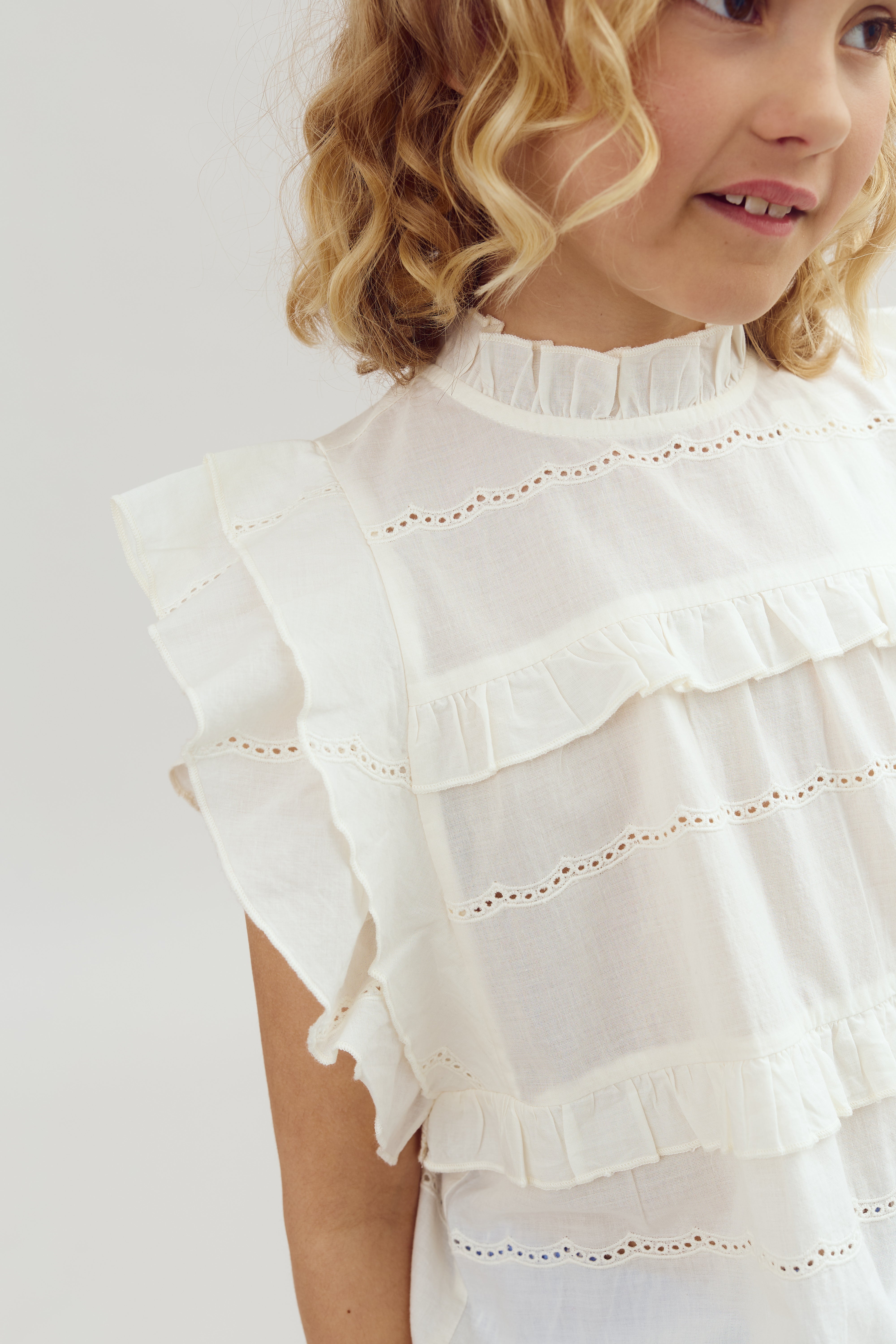 A child wearing a ruffled, white, embroidered blouse is partially turned away, displaying shoulder and face against a plain background. The blouse features lace details and layered edges.