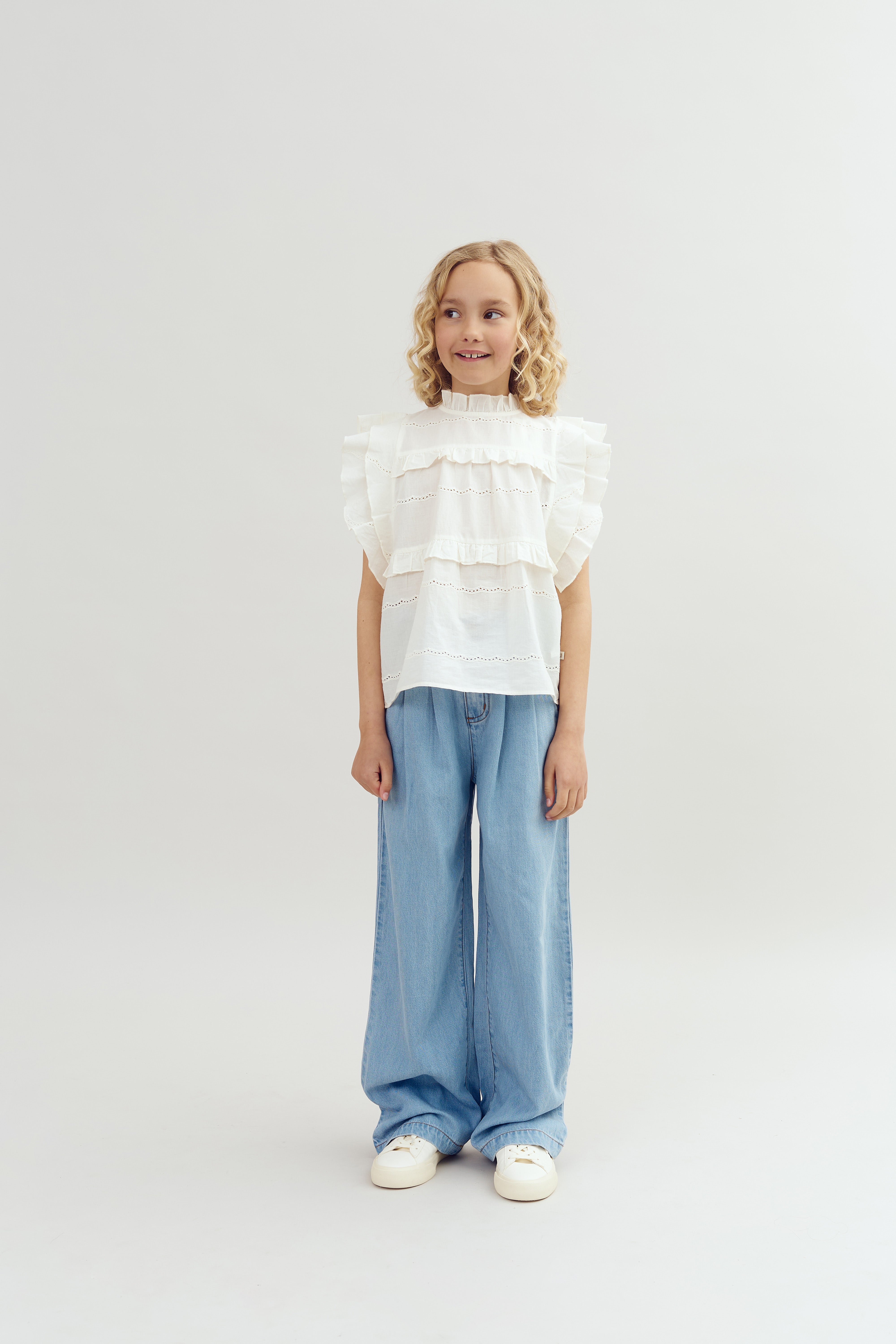 Barn står smilende iført en flæsebluse og brede denimjeans i et lyst studie med en neutral baggrund.