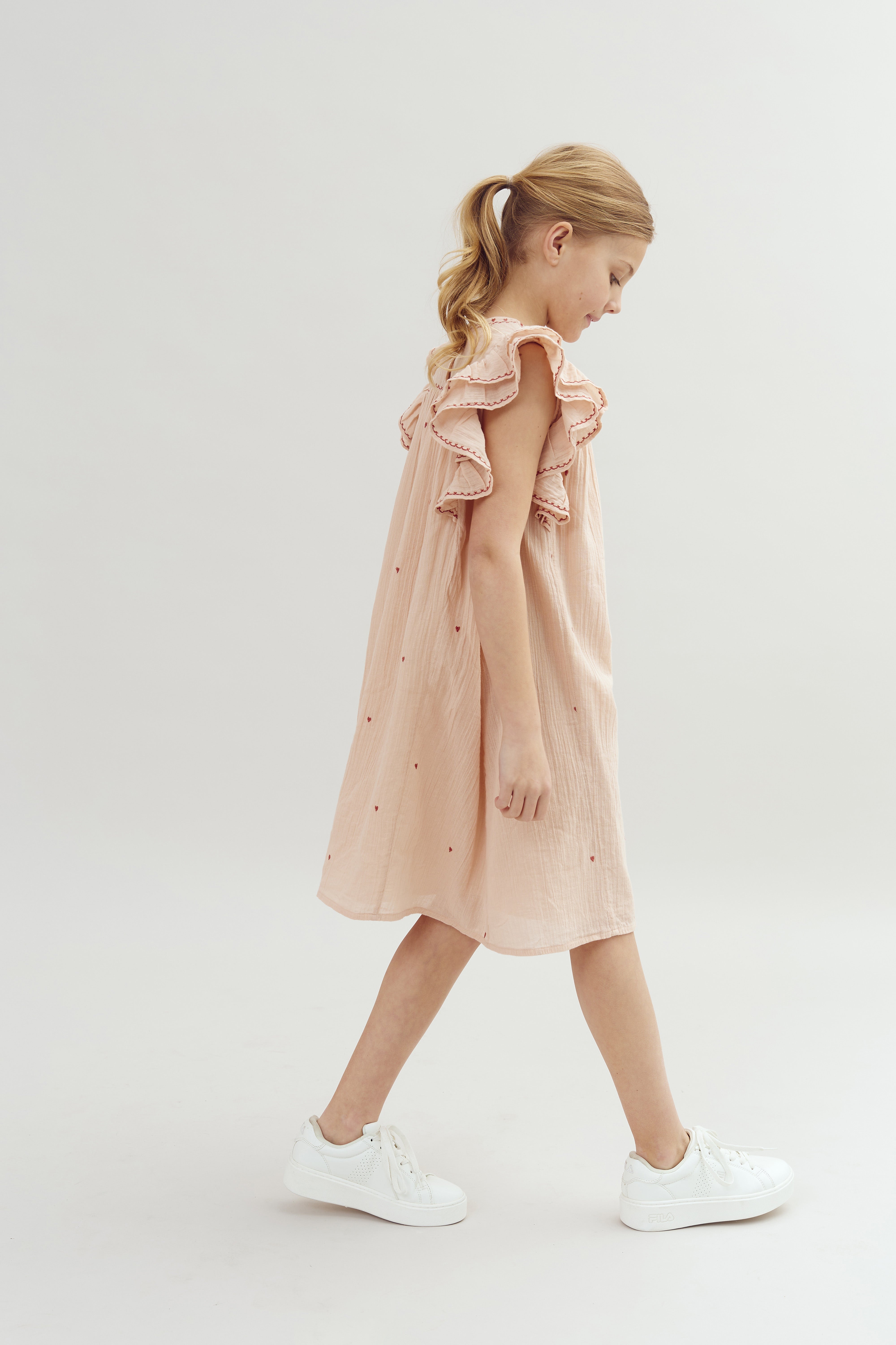 A young girl wearing a pink dress with ruffles and white sneakers walks forward in a minimal, light-colored studio setting.