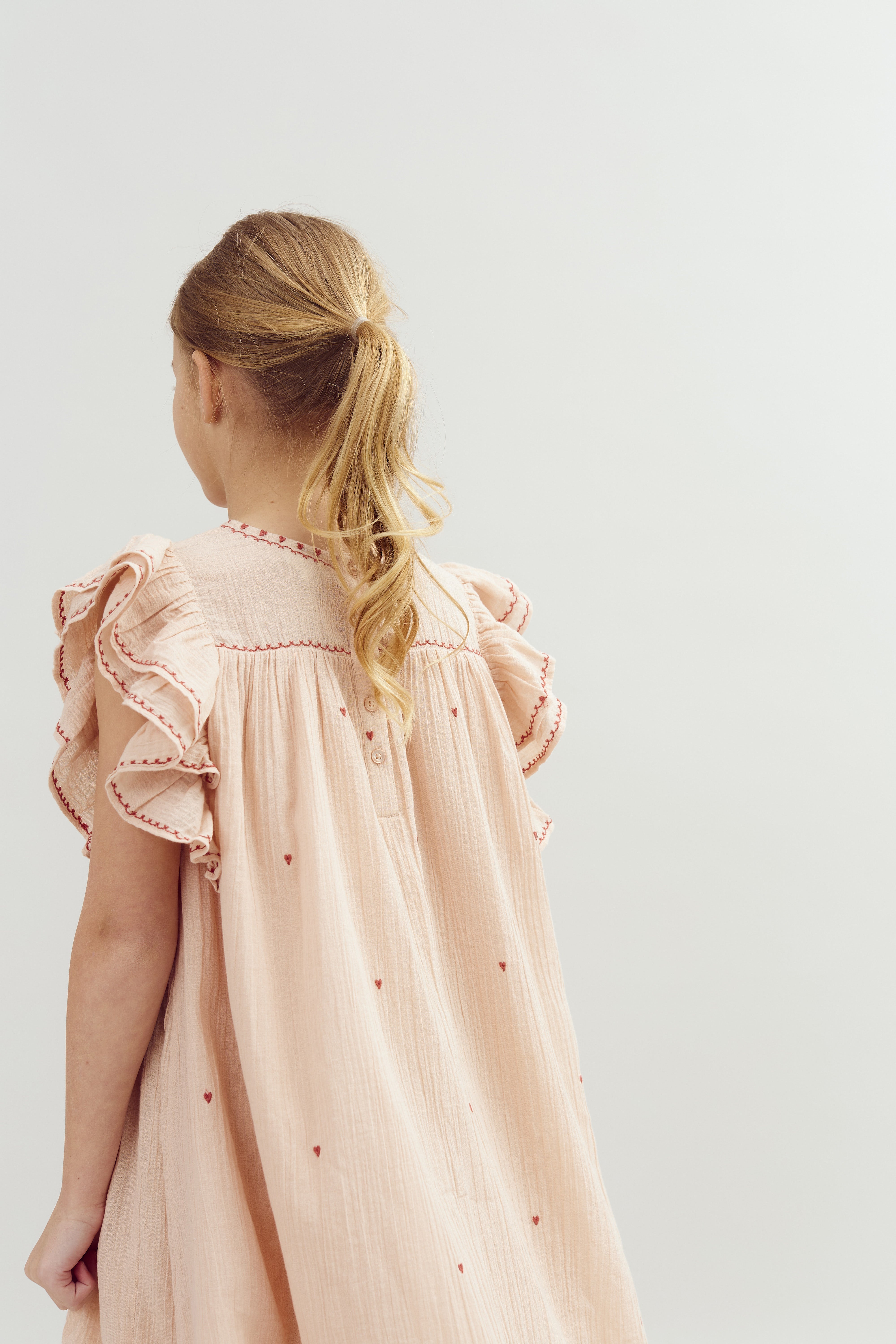 A young girl with blonde hair in a ponytail wearing a light pink dress with ruffled sleeves and red embroidery stands facing a plain neutral background.